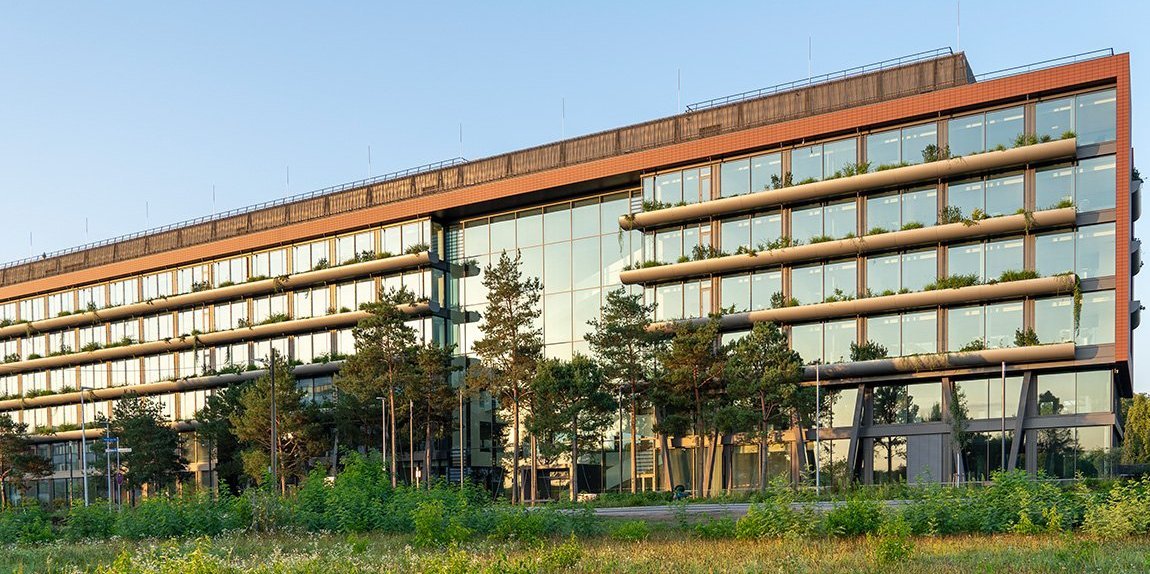 Neues Bürogebäude mit begrünter Fassade Glasfassade und Balkonen, umgeben von grüner Vegetation bei Sonnenuntergang.