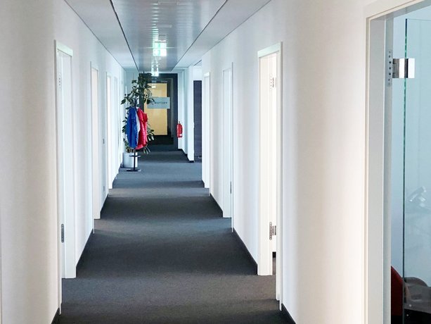 Büro Heller Büroflur mit weißen Wänden, mehreren Türen, Kleiderständer mit bunten Jacken und Pflanze im Hintergrund