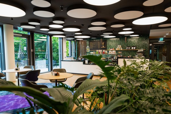 Kaffeebar mit stilvollem Interieur, runden Leuchten an der Decke, grüner Wand, großen Fenstern, gemütlichen Sitzplätzen und Pflanzen für ein einladendes Ambiente.