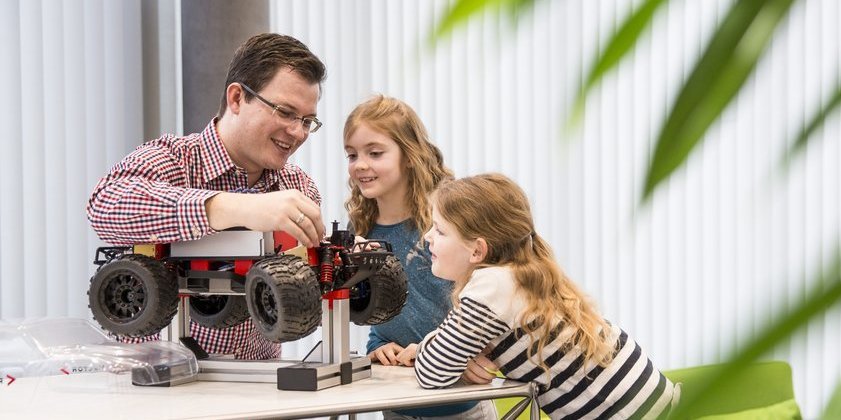 A colleague shows two schoolchildren a technical model car. They listen to his explanations with interest.