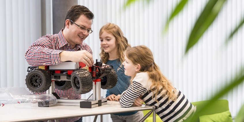 Ein Kollege zeigt zwei Schulkindern ein technisches Modellauto. Sie hören interessiert seinen Erklärungen zu.
