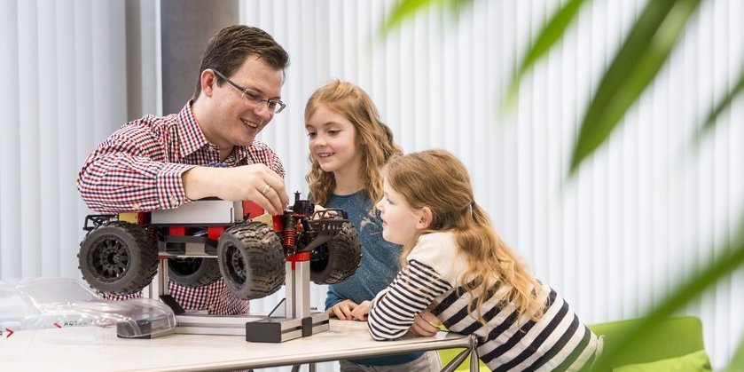 A colleague shows two schoolchildren a technical model car. They listen to his explanations with interest.