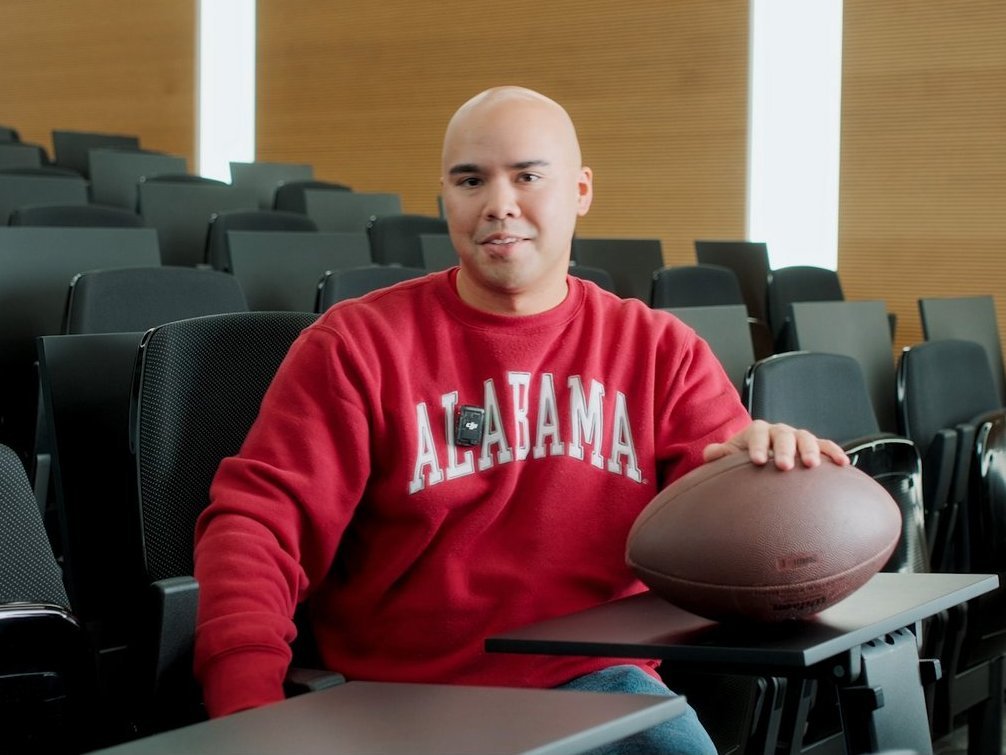 A Vector employee wearing a red sweatshirt is sitting in a modern auditorium. An American football is lying in front of him.