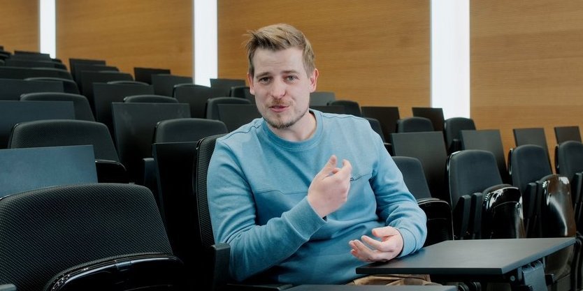 Vector-Mitarbeiter sitzt in einem Auditorium und gestikuliert, er trägt einen blauen Pullover.