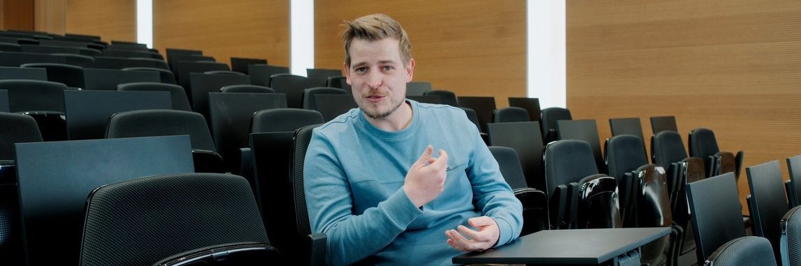 Vector-Mitarbeiter sitzt in einem Auditorium und gestikuliert, er trägt einen blauen Pullover.