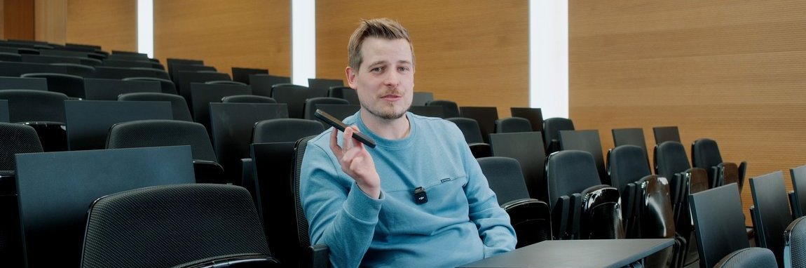 A Vector employee sits in an auditorium holding a smartphone in his hand. He is wearing a blue sweater.