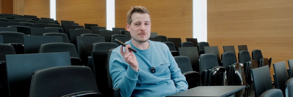 Vector-Mitarbeiter sitzt in einem Auditorium und hält ein Smartphone in der Hand, er trägt einen blauen Pullover.