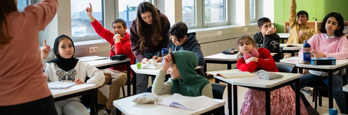 Schüler im Klassenzimmer, einige heben die Hand, Lehrerin vorne, zweite Lehrerin hilft einem Schüler – Szene aus dem Schulalltag.