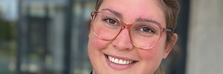 Portraitaufnahme einer Vector Mitarbeiterin. Sie lächelt vor einem unscharfen Bürohintergrund in die Kamera.