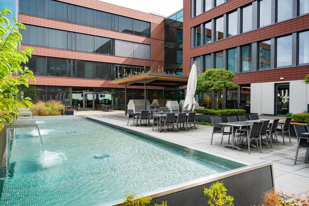 Innenhof moderner Bürogebäude mit Wasserfläche, Sitzgelegenheiten und Sonnenschirmen – urbaner Rückzugsort für Arbeit und Erholung im Freien
