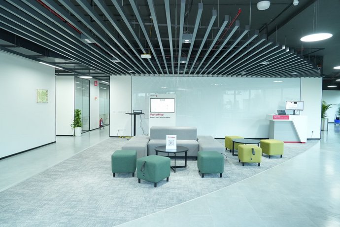 Modern communication zone with modular sofas and stools on a carpeted area in front of a large glass wall. The visible ceiling structure and integrated technology create a contemporary, high-quality office look.