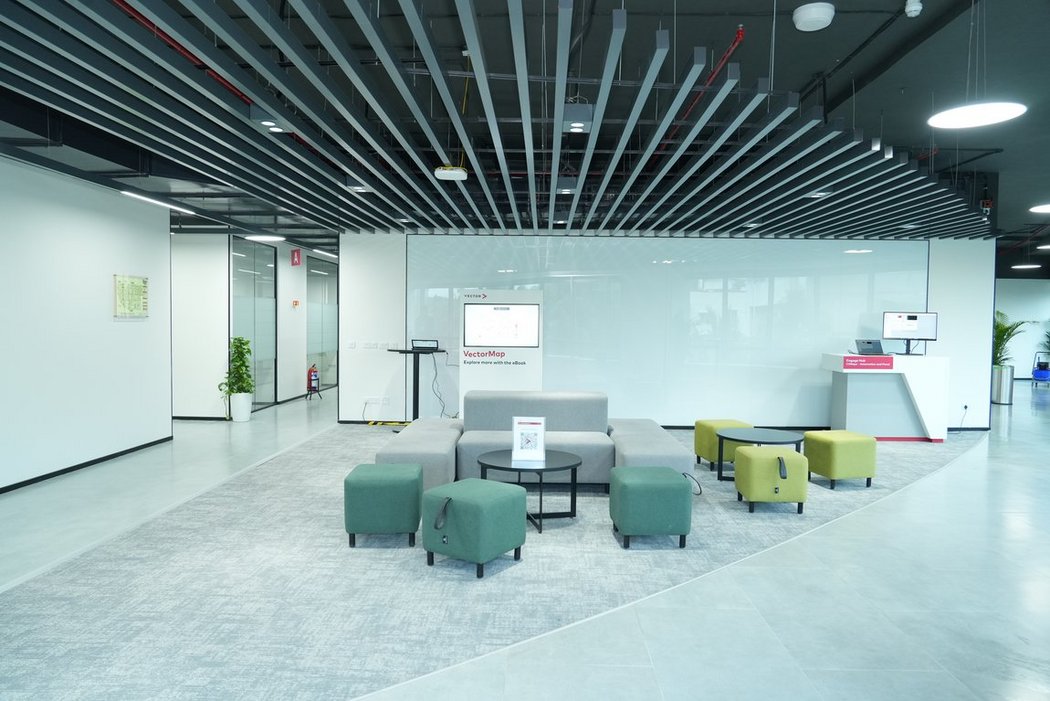 Modern communication zone with modular sofas and stools on a carpeted area in front of a large glass wall. The visible ceiling structure and integrated technology create a contemporary, high-quality office look.