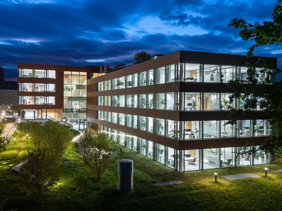 Bürogebäude bei Nacht mit beleuchteten Fenstern, Glasfassade, zeitgemäßem Design und grüner Umgebung