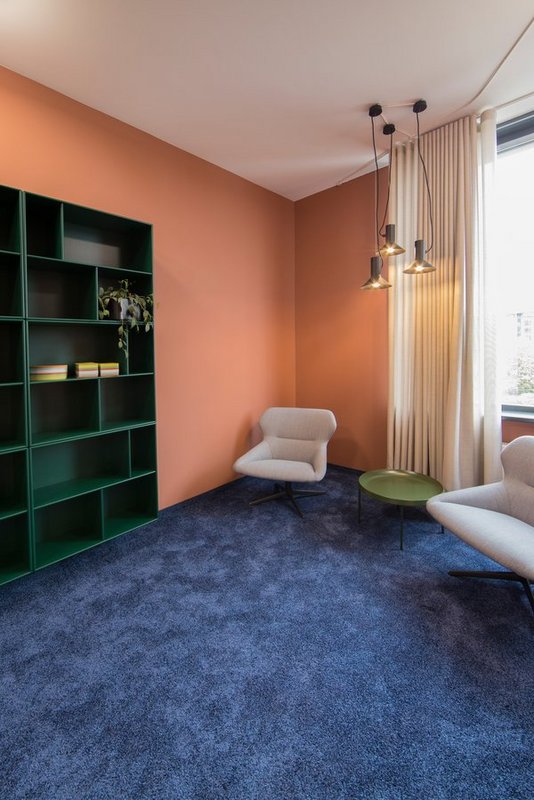 A room with a blue carpet, an orange wall, two armchairs and a shelf. 