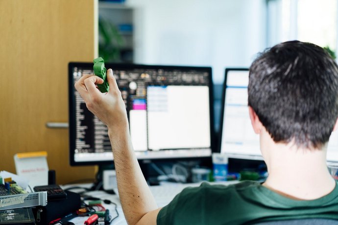 Person am Schreibtisch mit zwei Monitoren, hält grünes Objekt in der Hand – technisches Arbeiten mit Elektronik und digitalen Tools